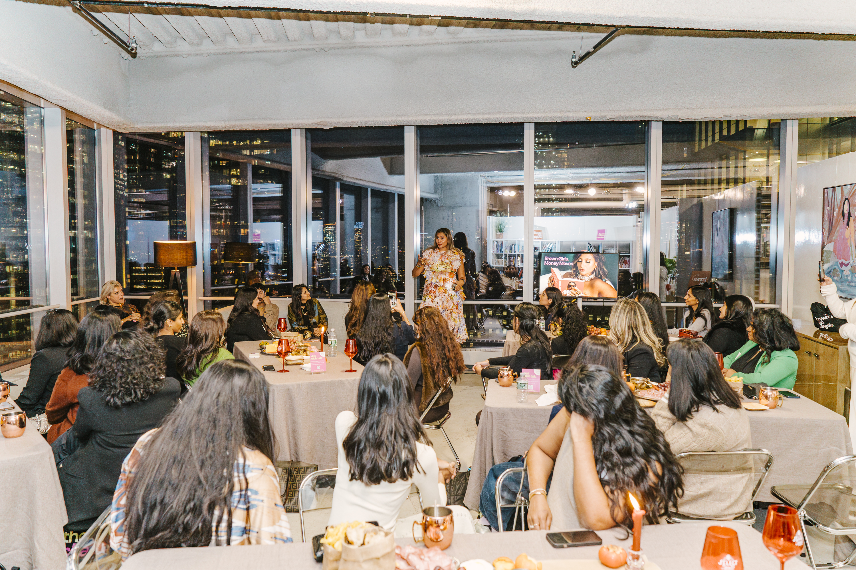 Bhargavi Varma addressing a room of senior South Asian women at a Brown Girl Angels community event in New York — the Brown Girls, Money Moves brand on screen, full NYC skyline behind her.